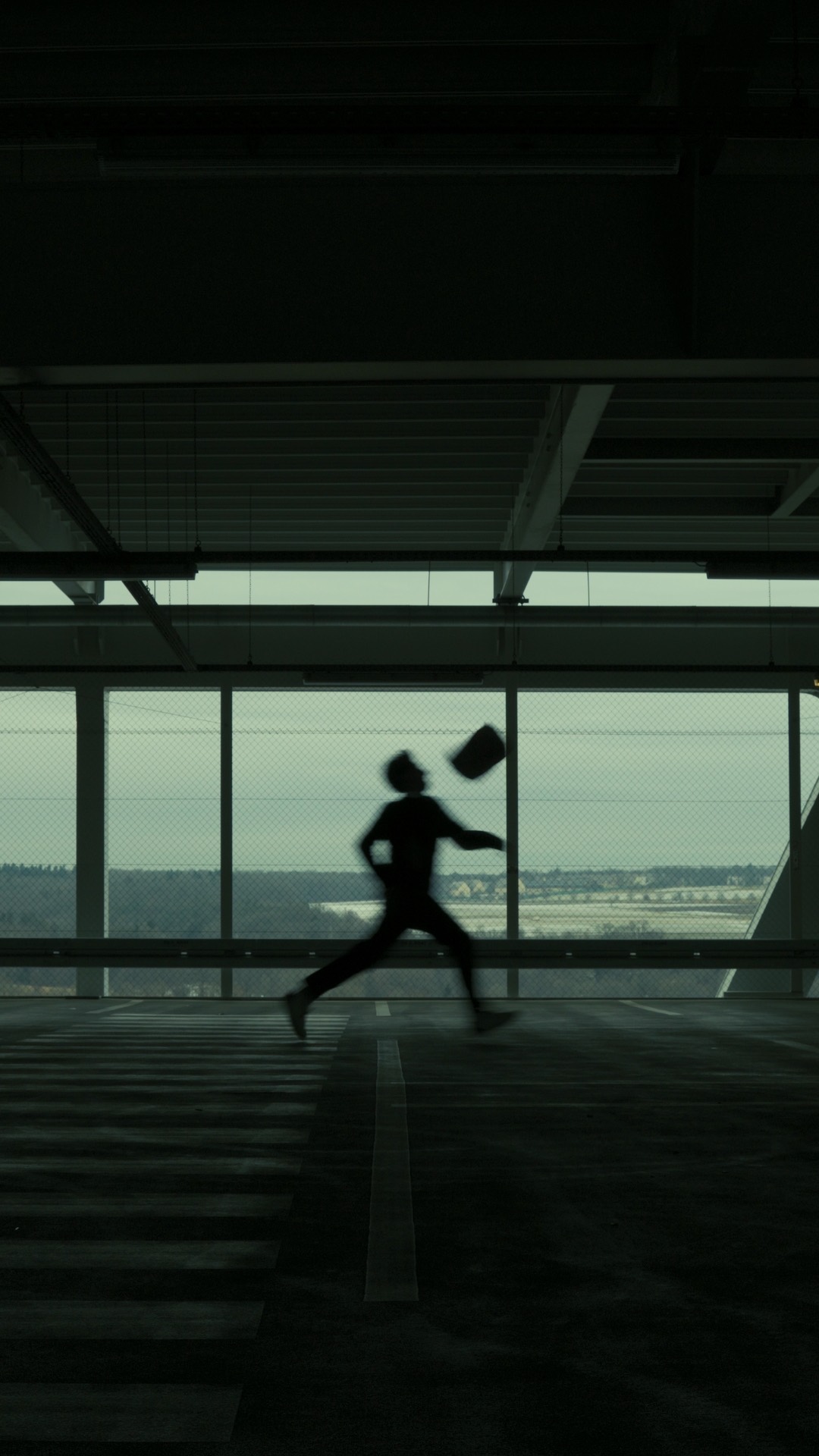 A figure in silhouette running through an empty concrete hall, a pale grey sky beyond the windows.